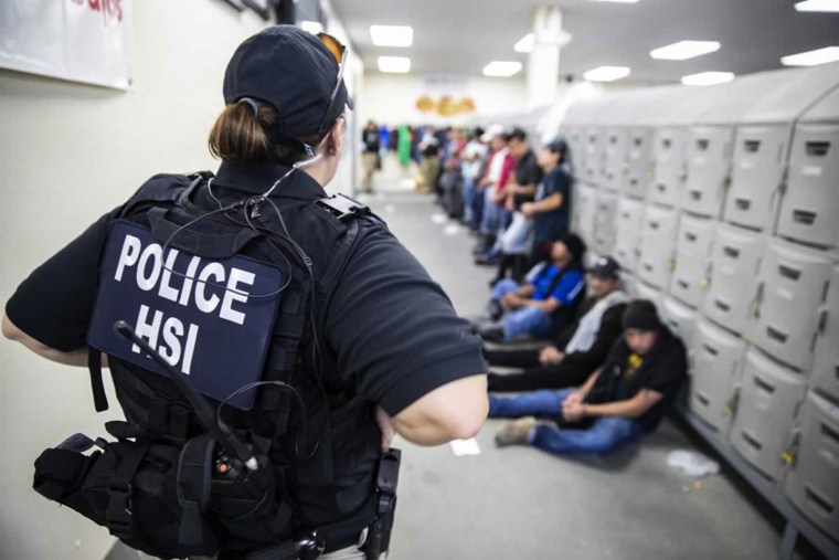 An officer stands in front of people seated and standing against lockers in a hallway