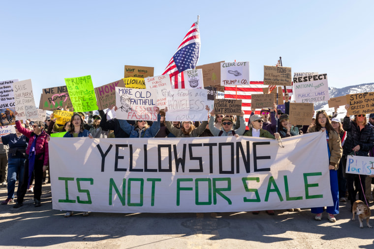 Protestors hold signs in protest outside, the banner in the front of the crowd reads "Yellowstone is not for sale"