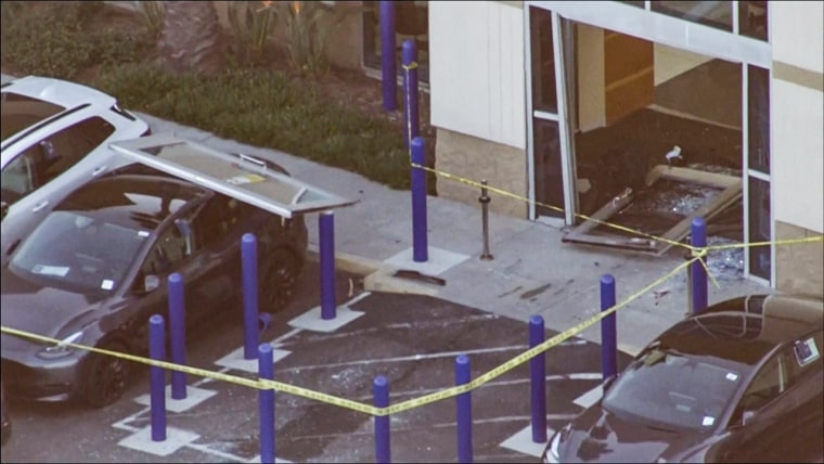 A broken glass door and car with a glass door landed on the trunk are seen outside a CarMax storefront