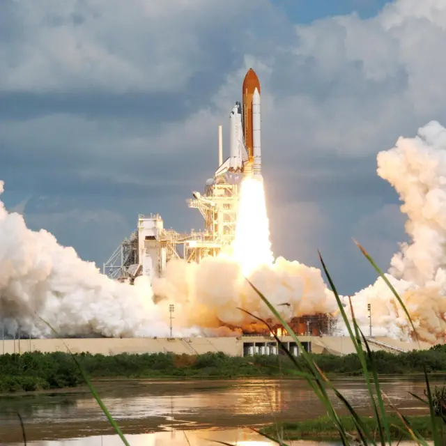 Space shuttle during launch from Cape Canaveral, Florida. The white ship is vertically attached to an orange rocket with large white thrusters. Flames shoot out of the motorbike and a large cloud of white smoke forms as the rocket prepares to take off. A lake appears in the foreground.