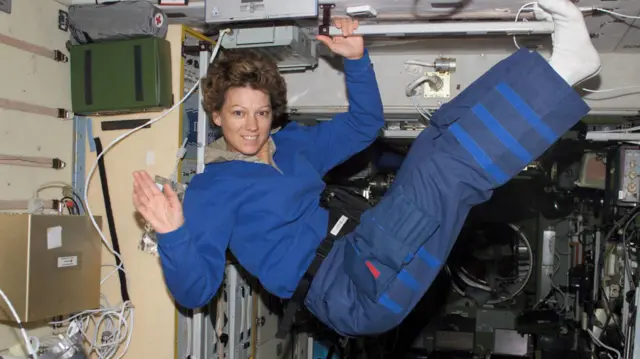 Eileen Collins floats inside a spaceship, smiling and waving at the camera, her legs high in the air. She wears a blue blouse and pants, with white socks, and holds a bar with her left hand. She is surrounded by wires and electronic equipment.