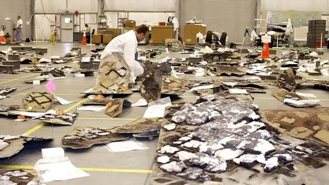 Debris from the space shuttle Columbia is laid out on the floor of a large warehouse. A man in a white coat leans over to inspect some of the material. There are boards and pieces of white foam, some of them burnt.