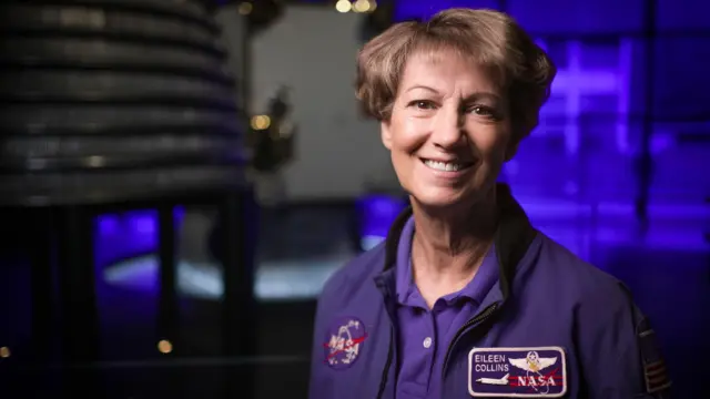 Eileen Collins wears a blue jacket with a NASA emblem on one side and a space shuttle patch with her name on the other. She has short brown hair and brown eyes. She is smiling at the camera. It's in the space gallery at the Science Museum in London and, in the background, out of focus, there is a rocket engine.
