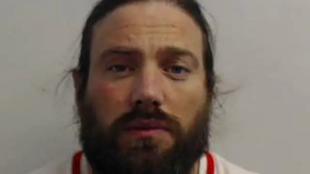 Police booking photo of Christopher Harkins. He faces the camera, with long hair tied back and a beard. 