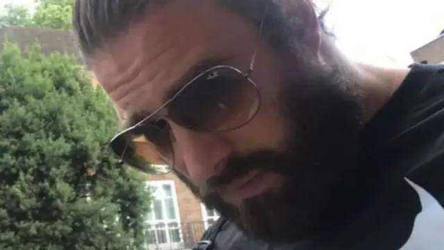 Photo of Christopher Harkins on his Tinder profile. It is outdoors, with trees and houses in the background. He has dark hair combed back and a full beard. He wears large sunglasses, with dark lenses and thin metal frames. Look down, towards the floor.