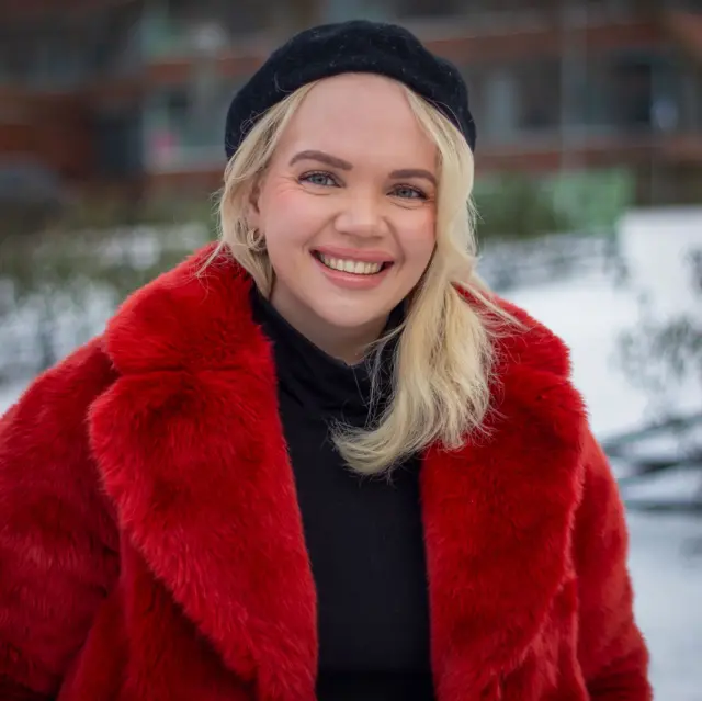 Meri-Tuuli Auer looking at the camera while wearing a fuzzy red coat and black beanie. Snow and trees can be seen in the background. 