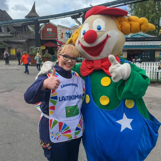 Meri-Tuuli Auer poses with a mascot dressed as a clown at an amusement park. They are giving a thumbs up to the camera.